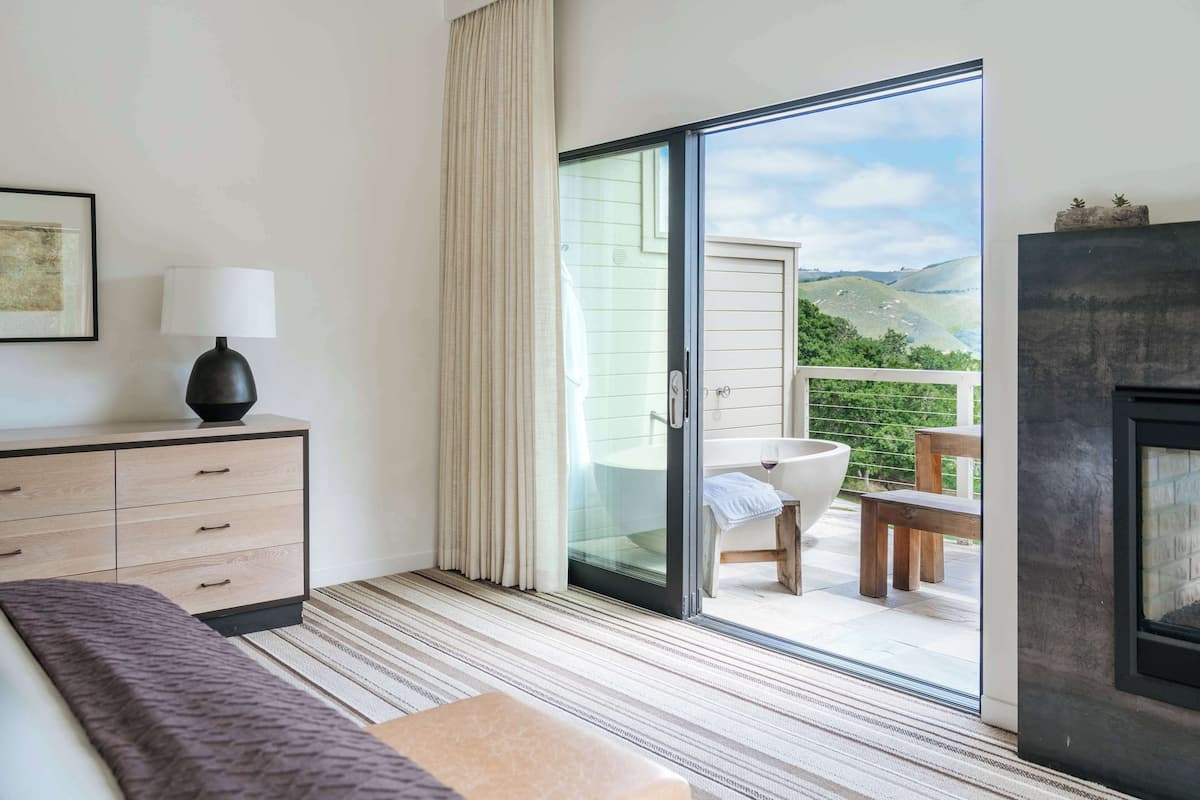A modern hotel room with a bed, a bedside table with a lamp, a bathroom with a sink and a chair, and a balcony with a view of the hills.