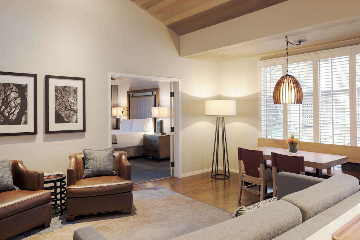 A hotel room with a living area featuring a sofa, armchairs, a dining table with chairs, a floor lamp, and artwork on the walls.
