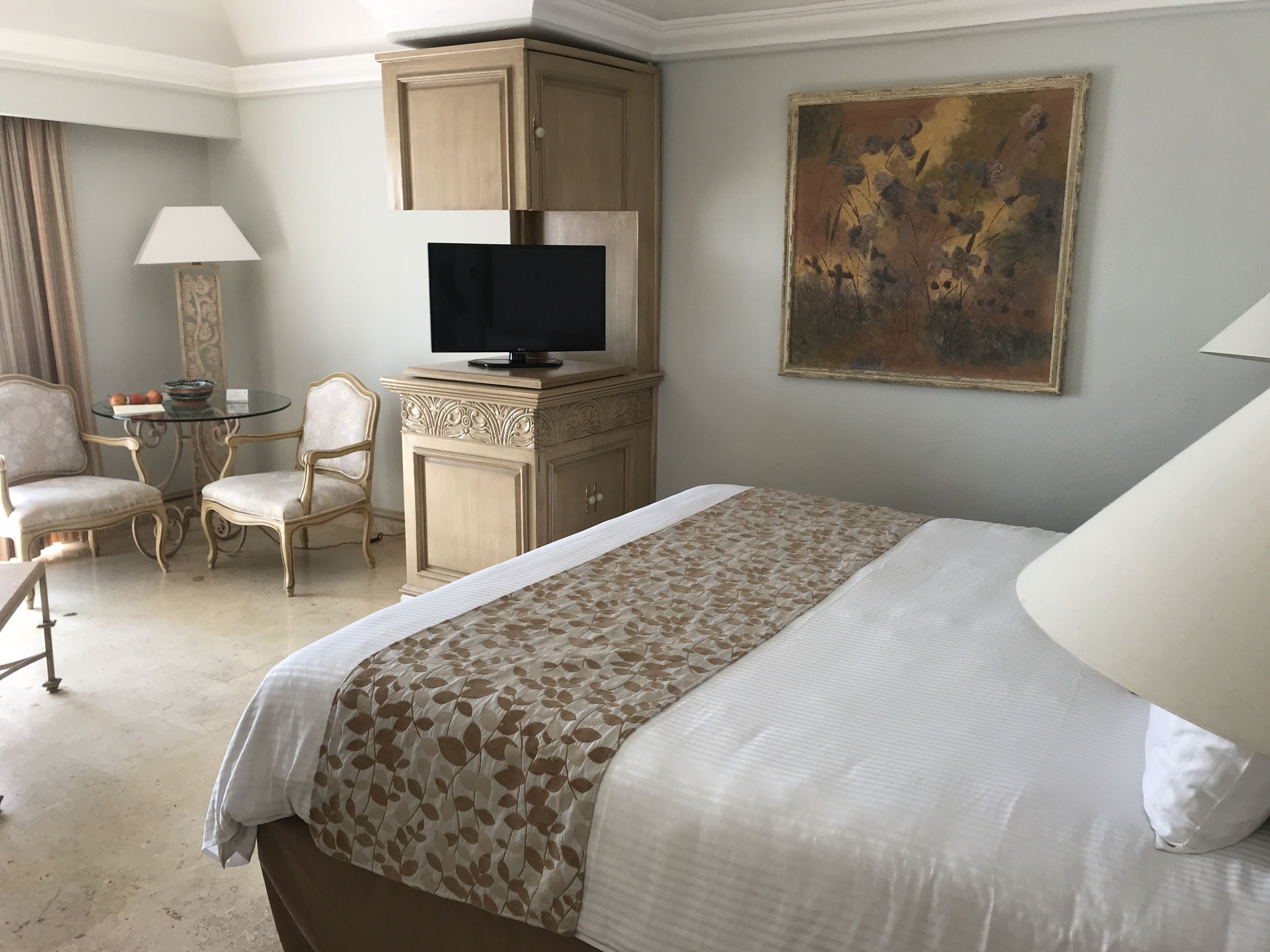 A hotel room with a large bed, a television on a wooden cabinet, a desk with a chair, and a painting on the wall.