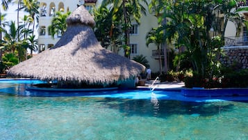 Un'area piscina con una struttura a tetto di paglia, circondata da palme e un edificio a più piani con balconi.