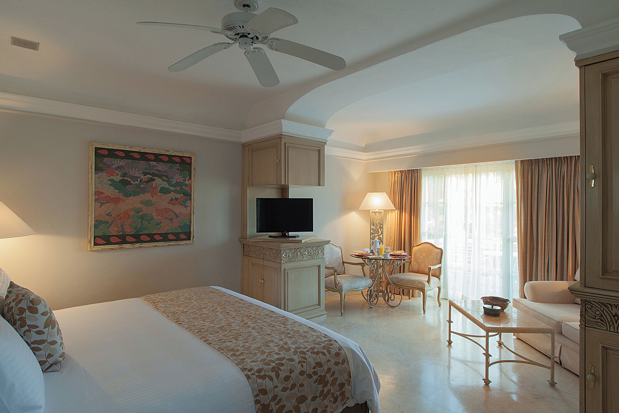 Une chambre d’hôtel avec un lit, un bureau avec un ordinateur, une télévision, un ventilateur de plafond et un tableau au mur.