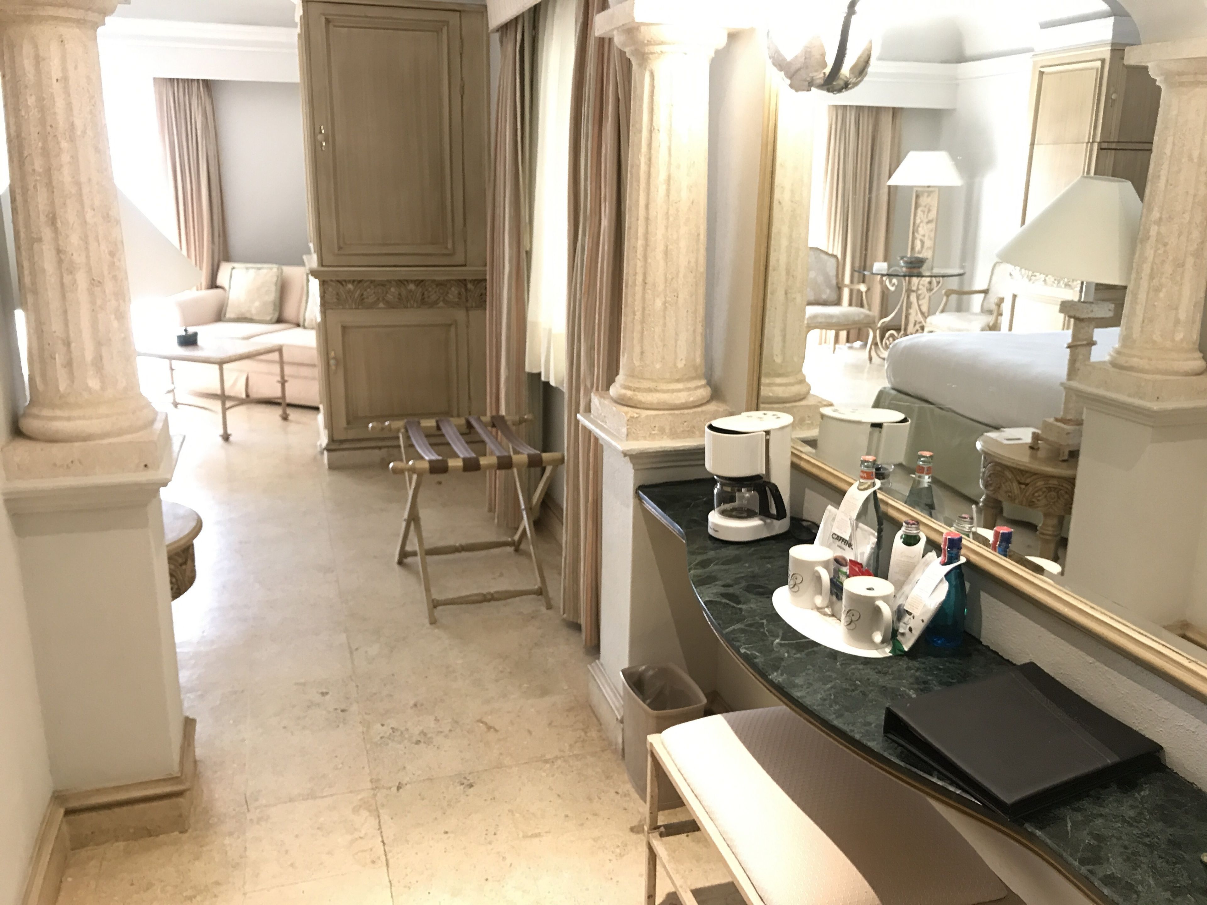 Habitación de hotel con cama, banca, cafetera, espejo y una mesita de noche con artículos de aseo.