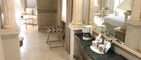 Habitación de hotel con cama, banca, cafetera, espejo y una mesita de noche con artículos de aseo.