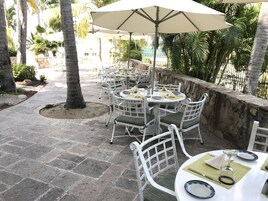 Un patio al aire libre con mesas redondas y sillas blancas, rodeado de palmeras y un muro de piedra.