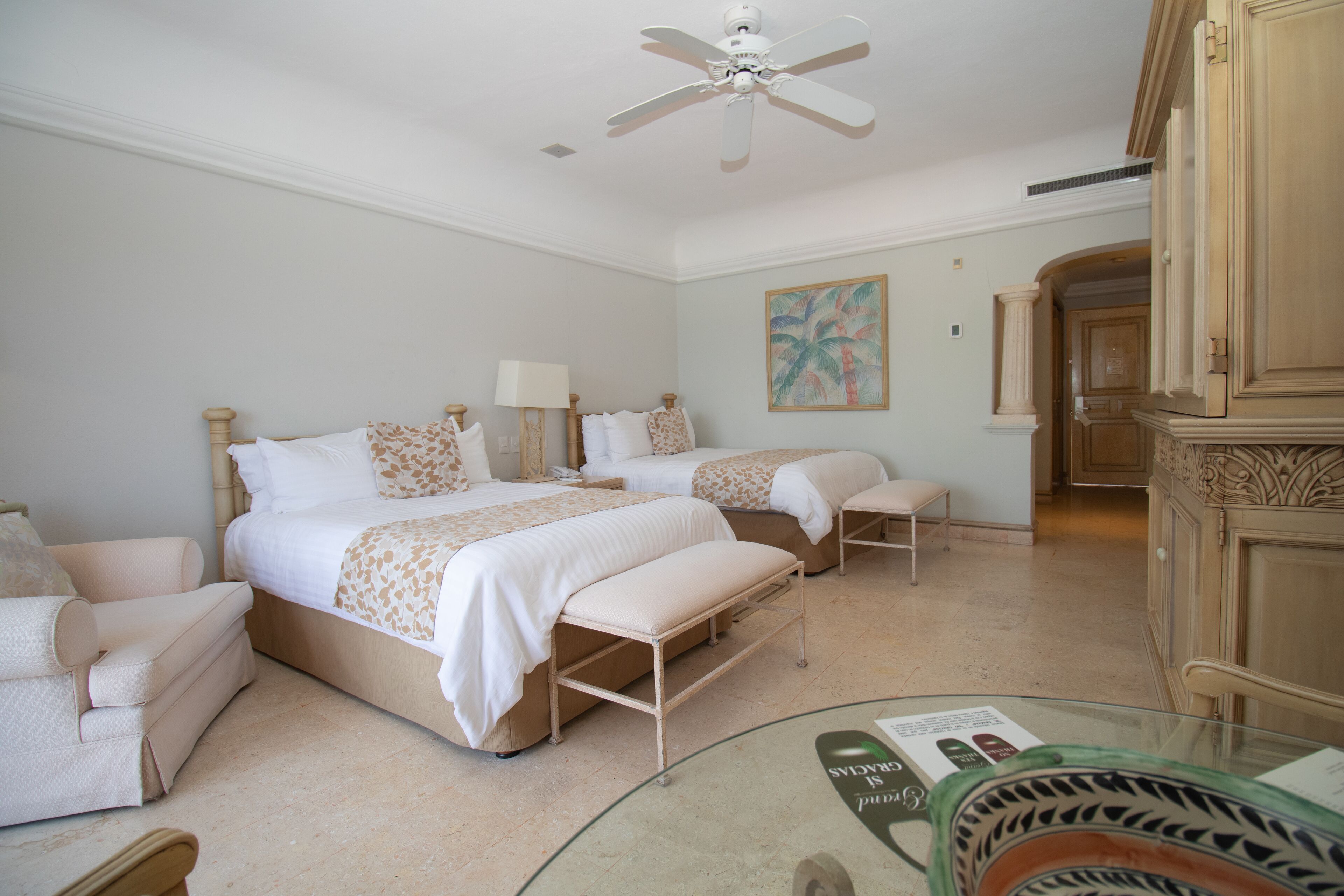 Une chambre d’hôtel avec deux lits, un canapé, une table basse en verre et un ventilateur de plafond.