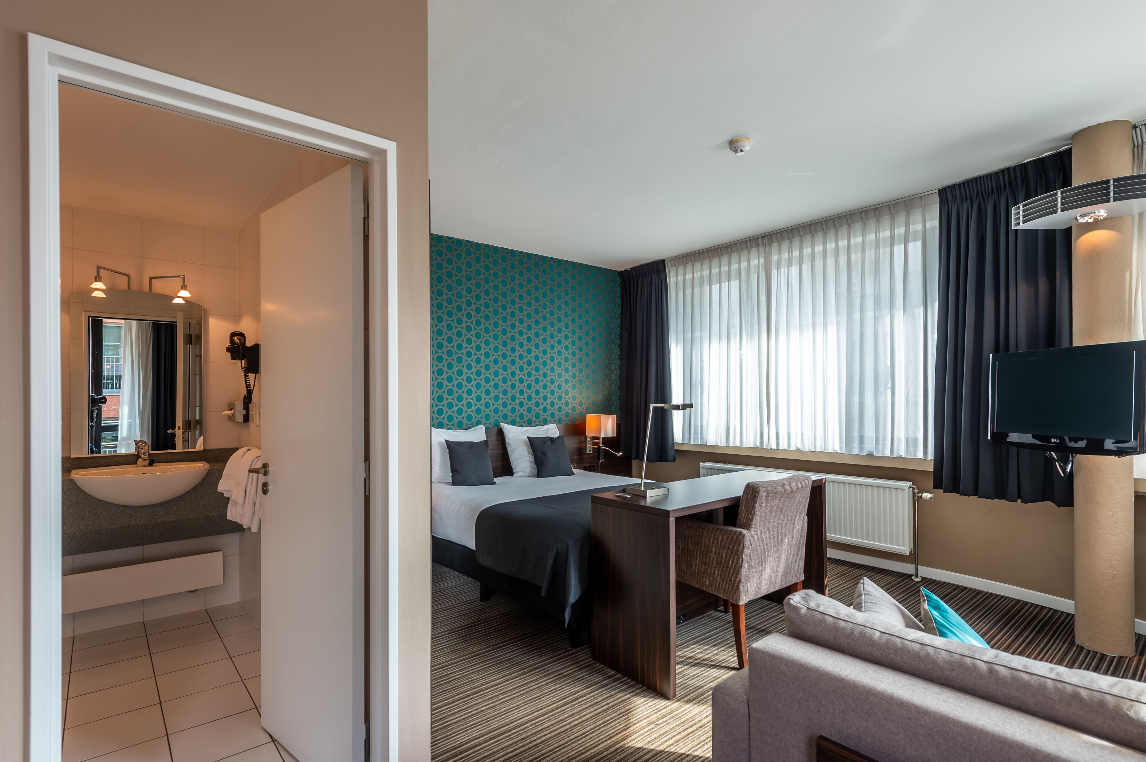 A hotel room with a large bed, a desk, a chair, a TV, and a bathroom with a sink and mirror.