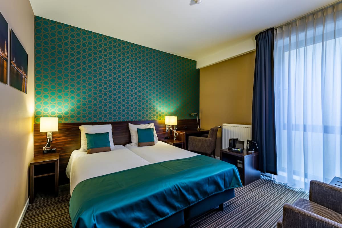 A hotel room with a large bed, two bedside tables with lamps, a desk with a chair, and a window with curtains.