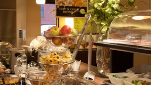 Un bufé con diferentes opciones de comida, entre ellas frutas y quesos, en una sala bien iluminada con luces colgantes y un cartel que indica bebidas calientes y frías.