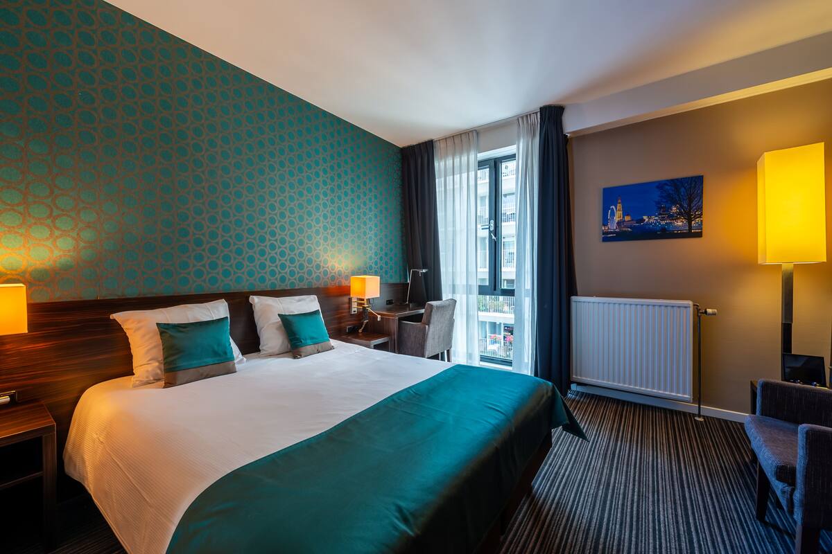 A hotel room with a large bed, two bedside tables with lamps, a desk, a chair, a view of the city through a window, and a painting on the wall.