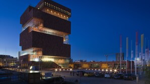 Un edificio moderno con varios pisos, ventanas amplias y un estacionamiento con coches y banderas.