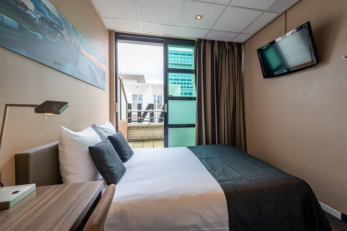 A hotel room with a bed, a desk, a lamp, a TV, and a painting on the wall.