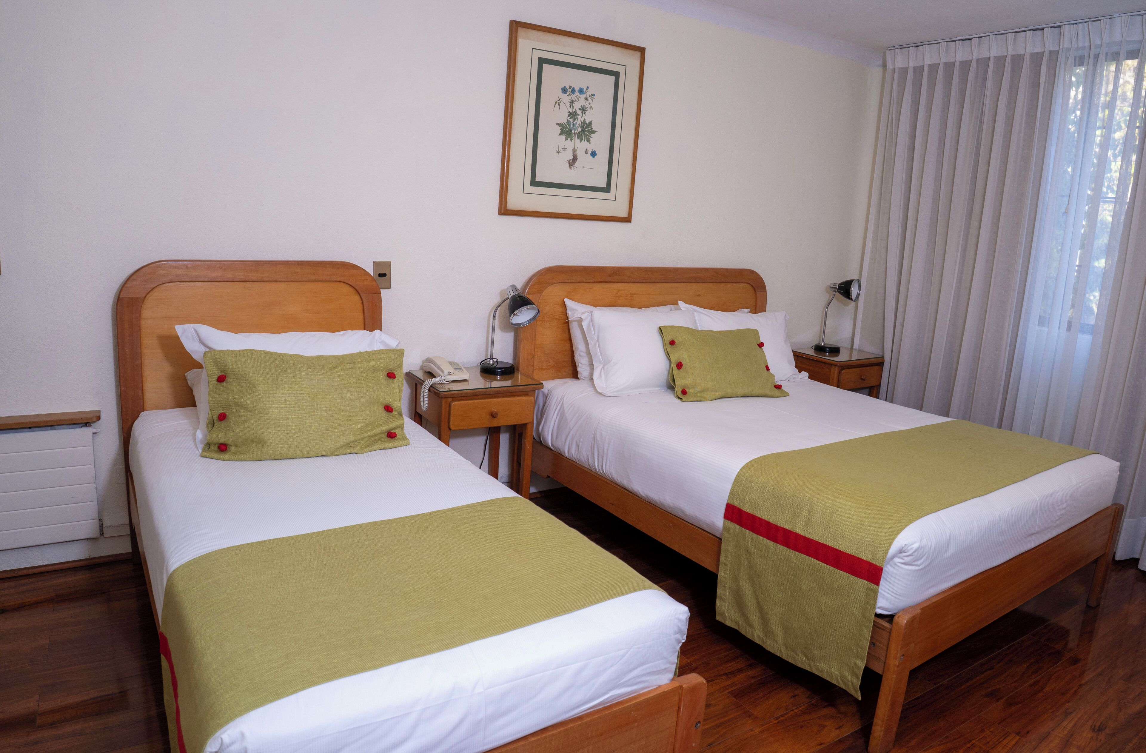 Habitación de hotel con dos camas individuales, cabeceros de madera, ropa de cama blanca, una mesita de noche con lámpara e impresión botánica enmarcada en la pared.
