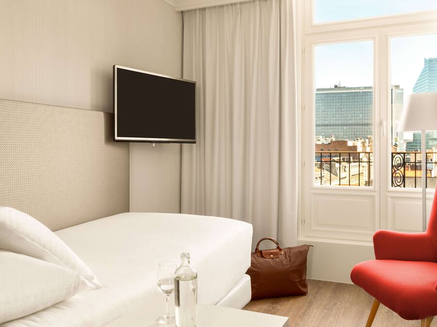 Habitación de hotel con cama, televisor, ventana con vista a edificios, una silla roja y un bolso marrón.