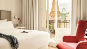 Habitación de hotel con cama, un sillón rojo, una mesita con un jarrón de flores y vista a un edificio a través de una puerta de cristal.