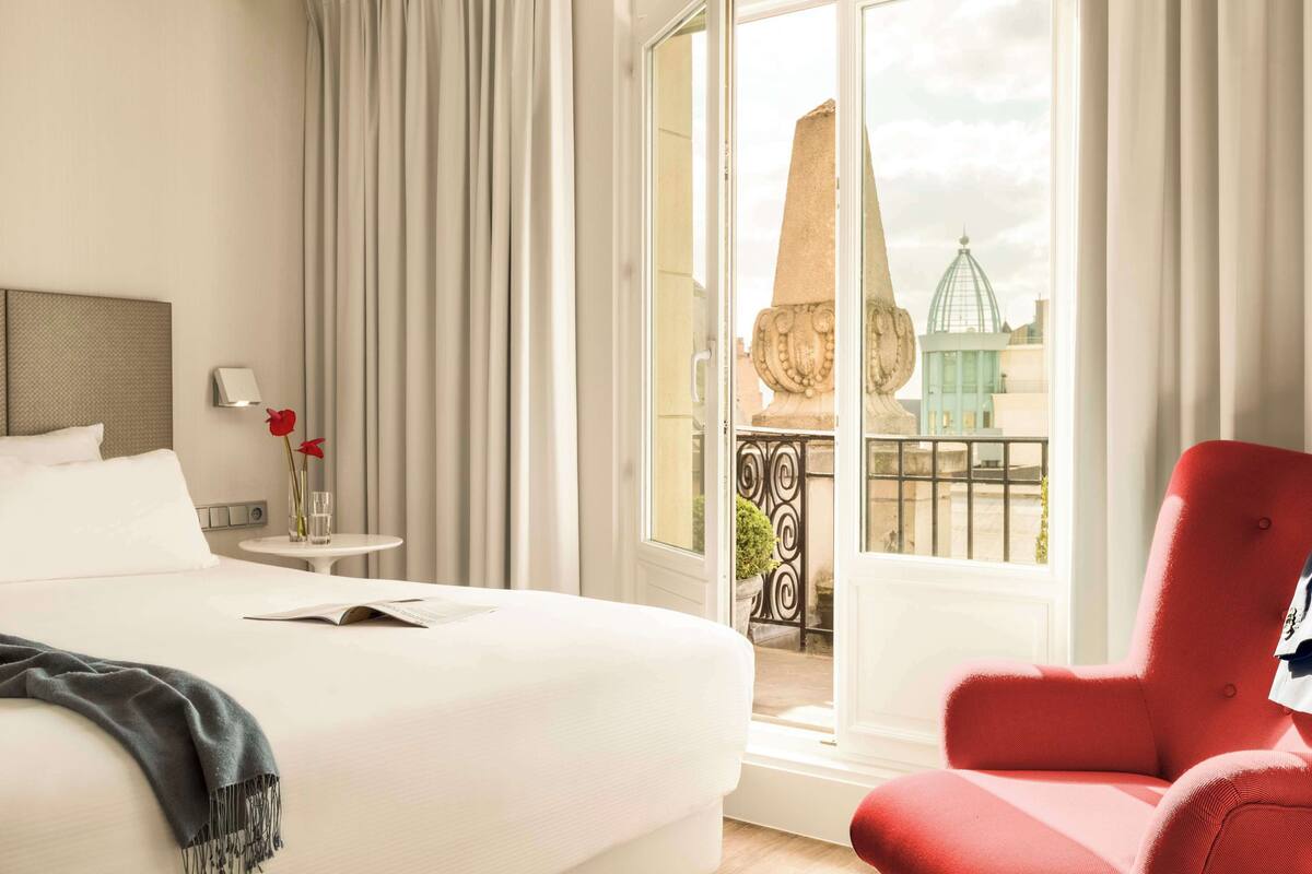 A hotel room with a bed, a red armchair, a small table with a vase of flowers, and a view of a building through a glass door.