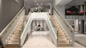 Un moderno vestíbulo de hotel con una escalera central, barandales de vidrio y un área de descanso con mesas y sillas.