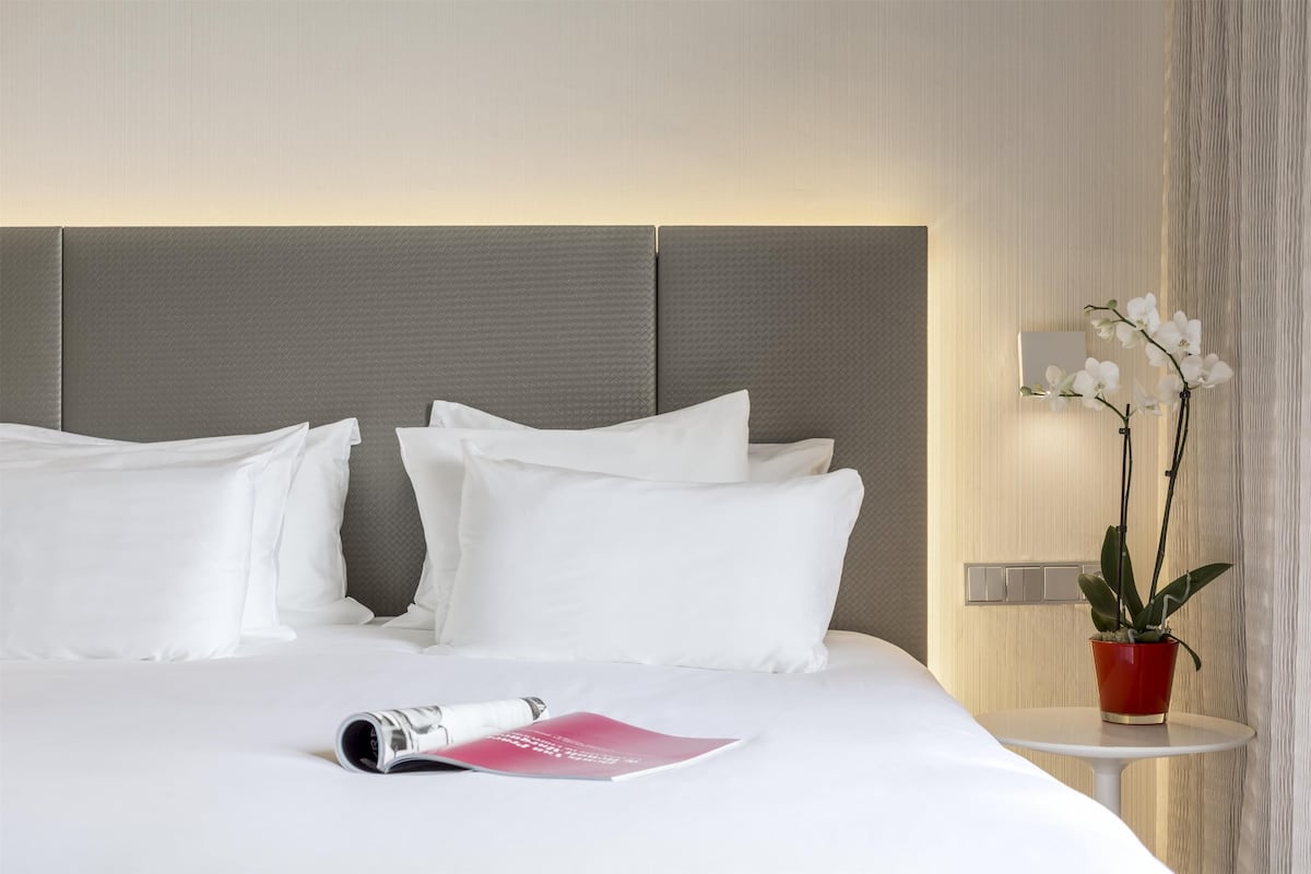 A neatly made bed with white linens and pillows, a small table with a red pot of flowers, and a wall-mounted light fixture.