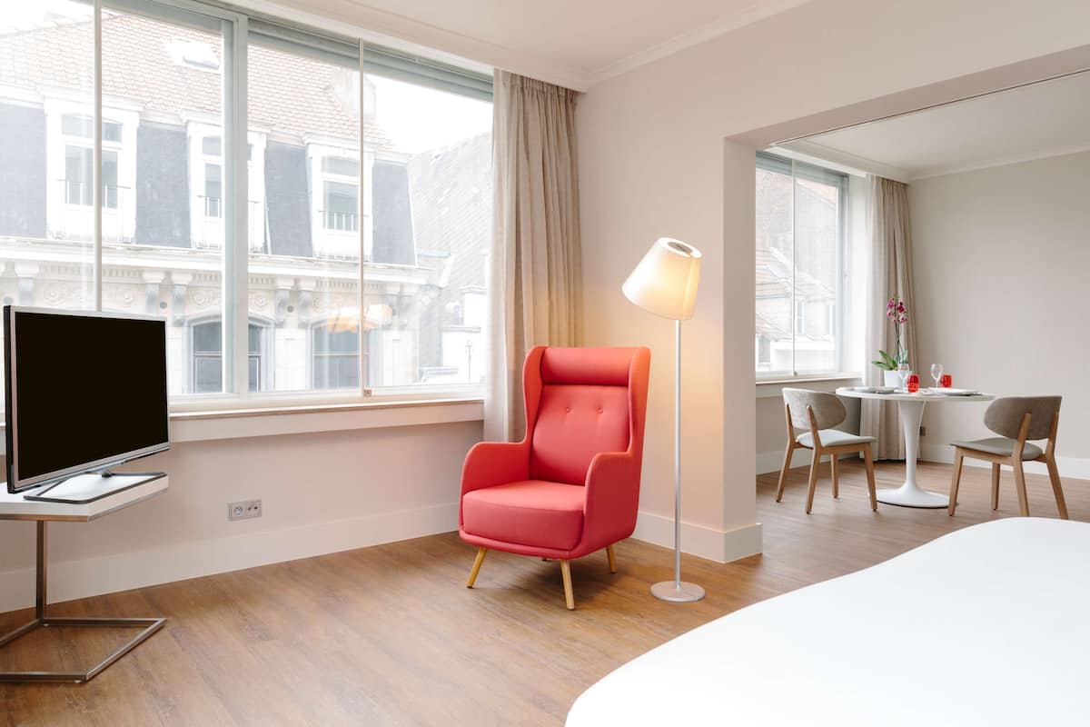 A hotel room with a flat-screen TV, a red armchair, a floor lamp, a round dining table with chairs, and large windows with curtains.