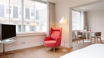 A hotel room with a flat-screen TV, a red armchair, a floor lamp, a round dining table with chairs, and large windows with curtains.