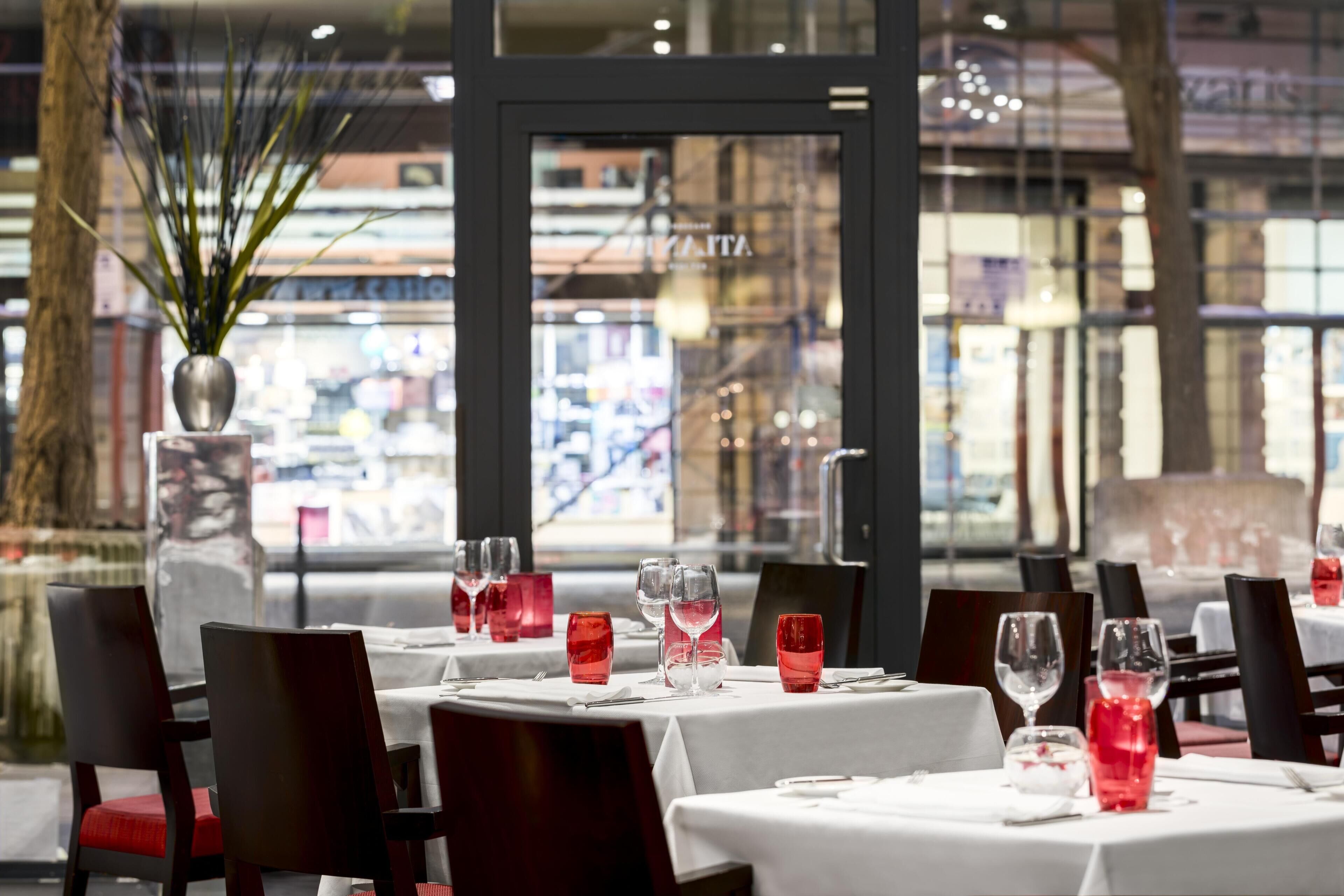 A restaurant interior with round tables set for dining, red glassware, and a large potted plant.