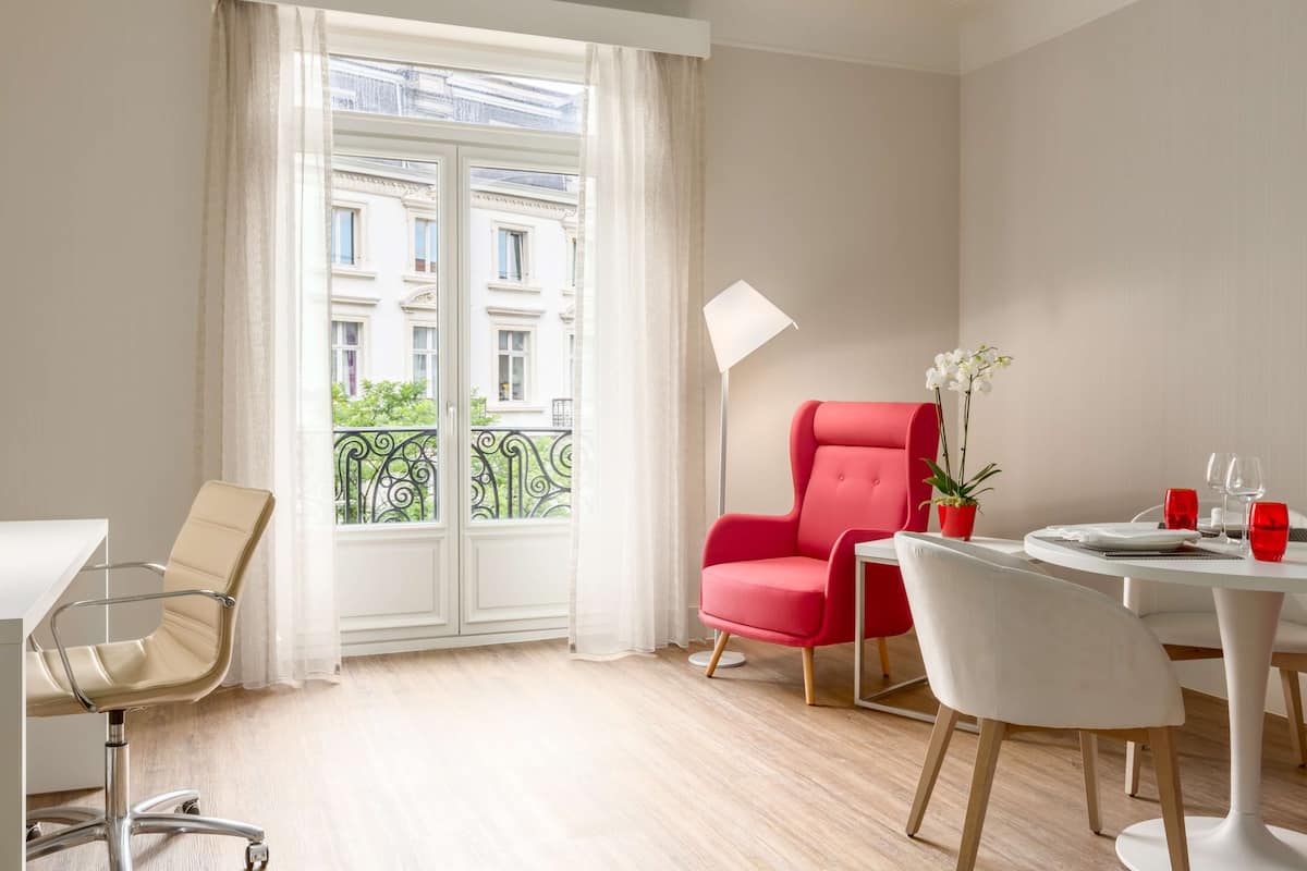 A modern hotel room with a dining area, a red chair, a white chair, a table, and a potted plant.