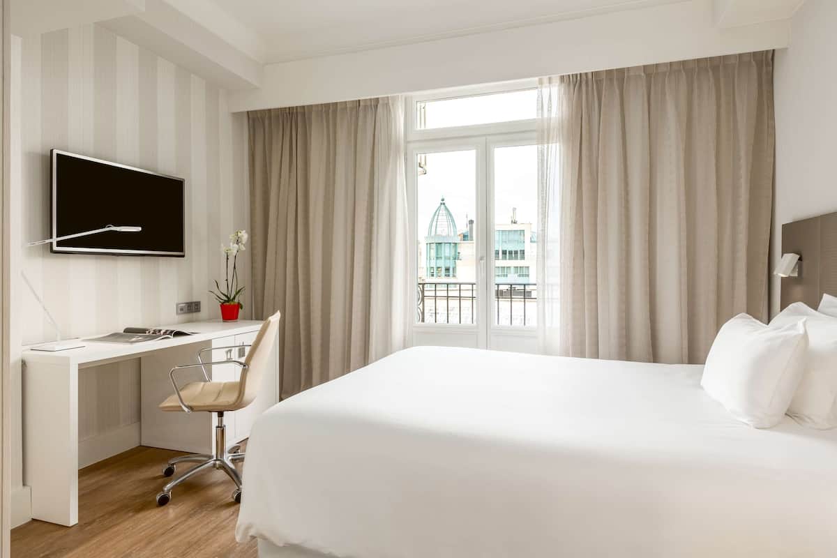 A hotel room with a bed, a desk, a chair, a TV, and a window with curtains.