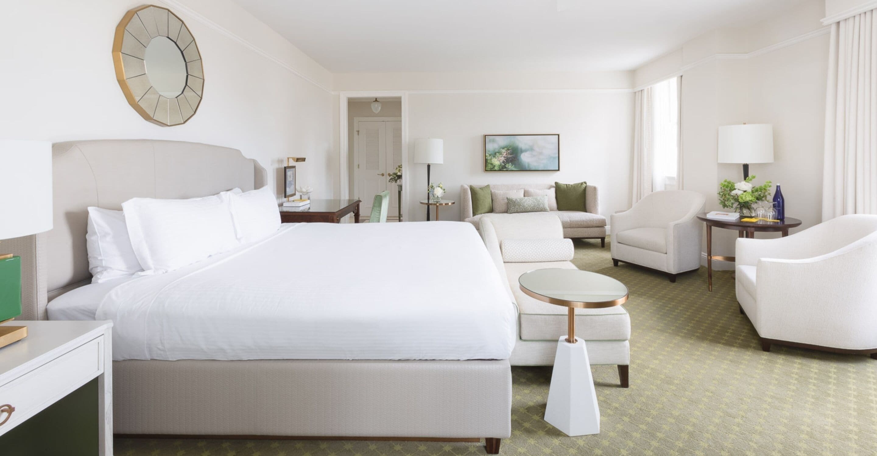 A hotel room with a large bed, a round coffee table, a sofa, and a chair. There is a painting on the wall and a clock above the bed.