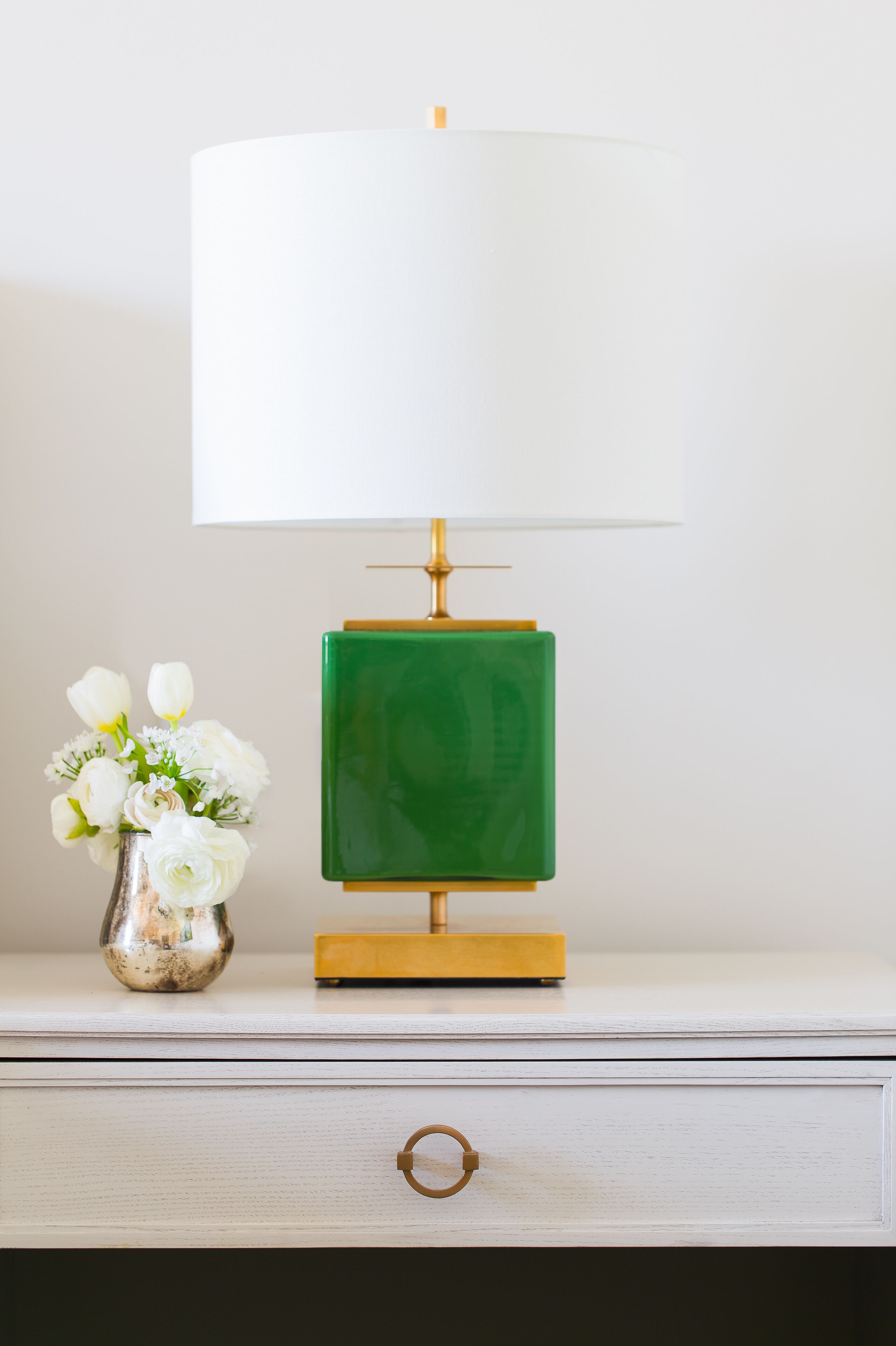 Ein Nachttisch mit einer grün-goldenen Lampe, einer weißen Vase mit Blumen und einer weißen Schublade mit Messingring.
