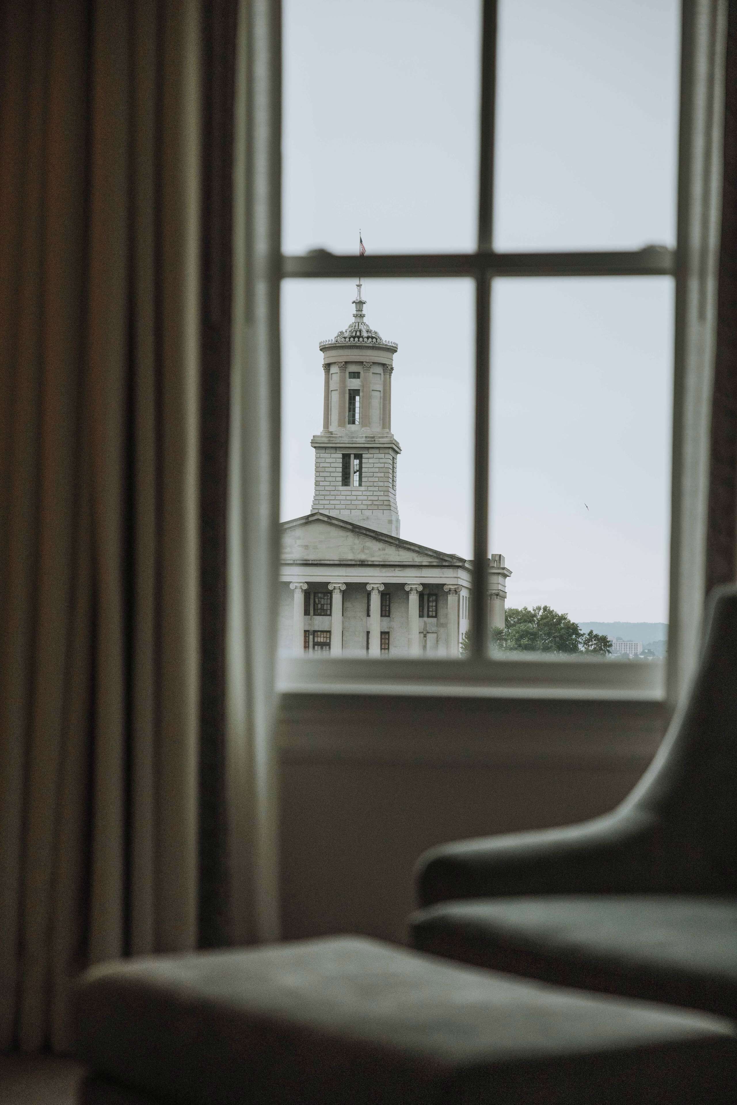 A view through a window of a building with a prominent tower and columns, visible curtains, and a chair in the foreground.