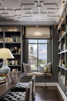 A room with a large window, bookshelves, a table with a lamp, and a bench.