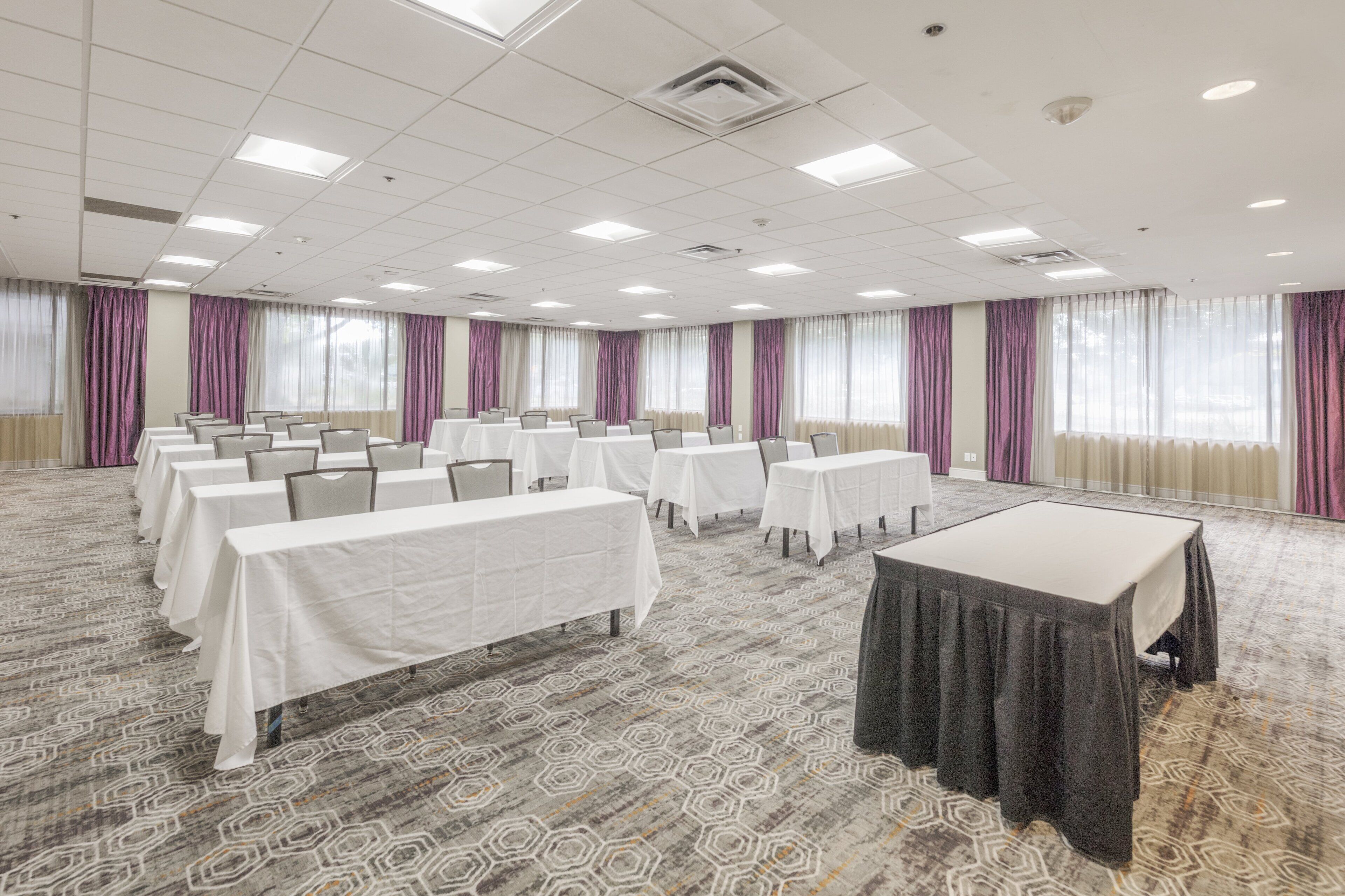 Une salle de conférence dotée de plusieurs tables et chaises, de grandes fenêtres avec des rideaux et d’un tapis à motifs.