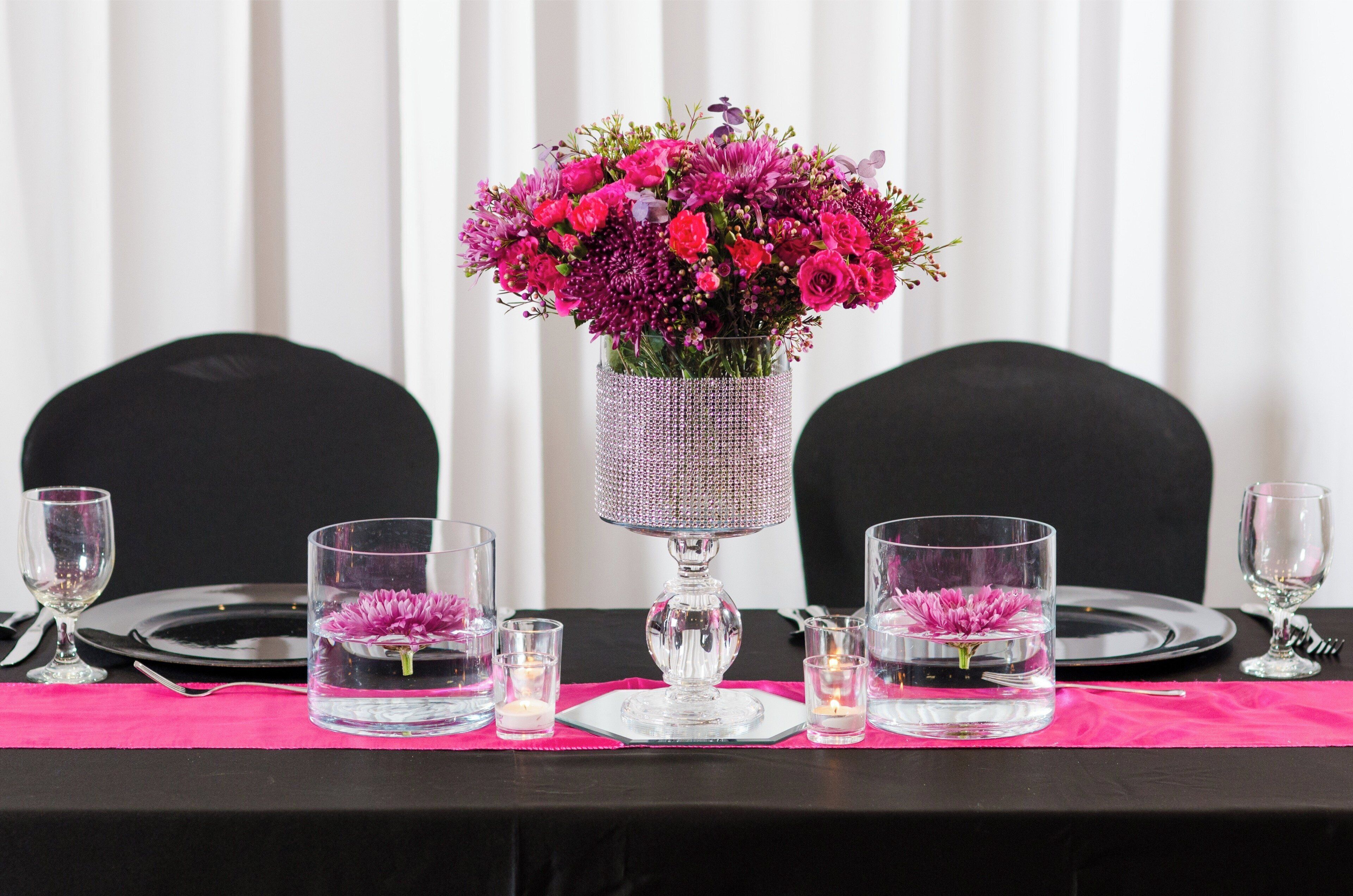 A table set for dining with a centerpiece of pink flowers in a crystal vase, clear glasses, and black tableware.