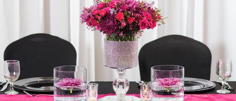 Une table dressée pour le dîner, ornée d’un centre de table composé de fleurs roses dans un vase en cristal, de verres transparents et de vaisselle noire.