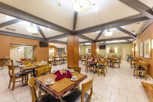 A spacious dining area with multiple wooden tables and chairs, high ceilings with exposed beams, and a television mounted on the wall.