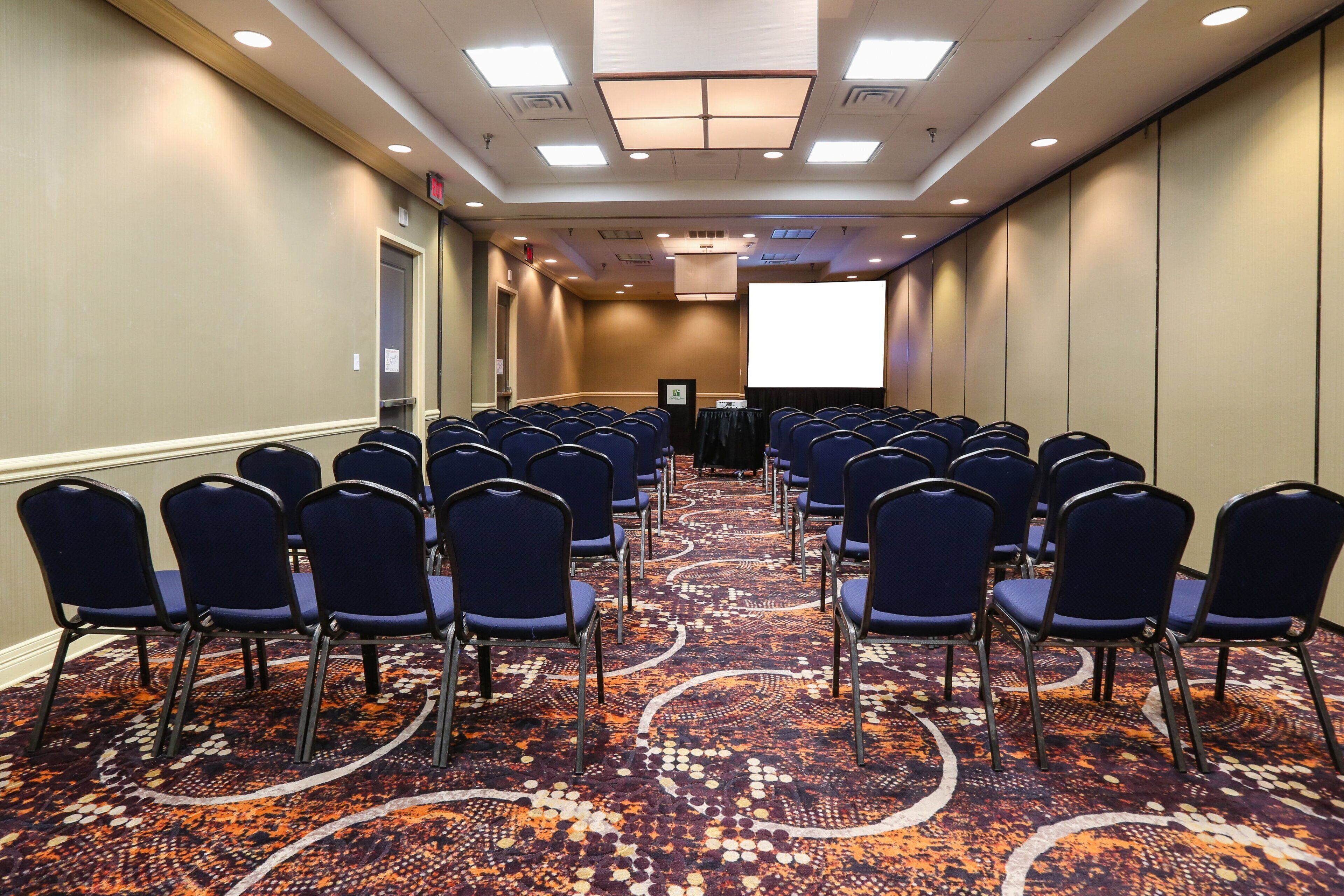 A conference room with rows of chairs facing a projection screen, carpeted floor, and a door on the left.