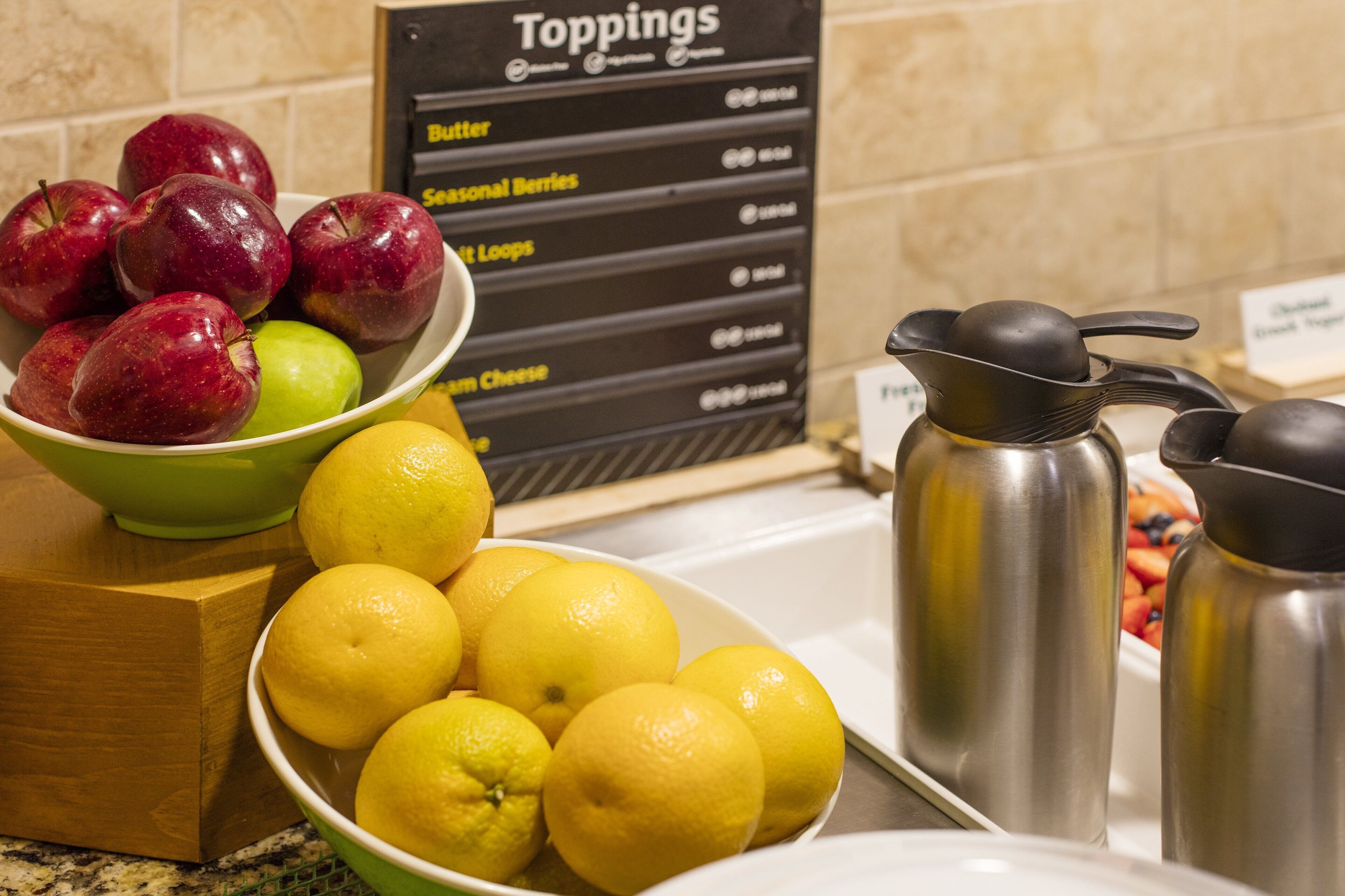 A kitchen counter with a bowl of apples and lemons, a sign listing toppings, and stainless steel containers.