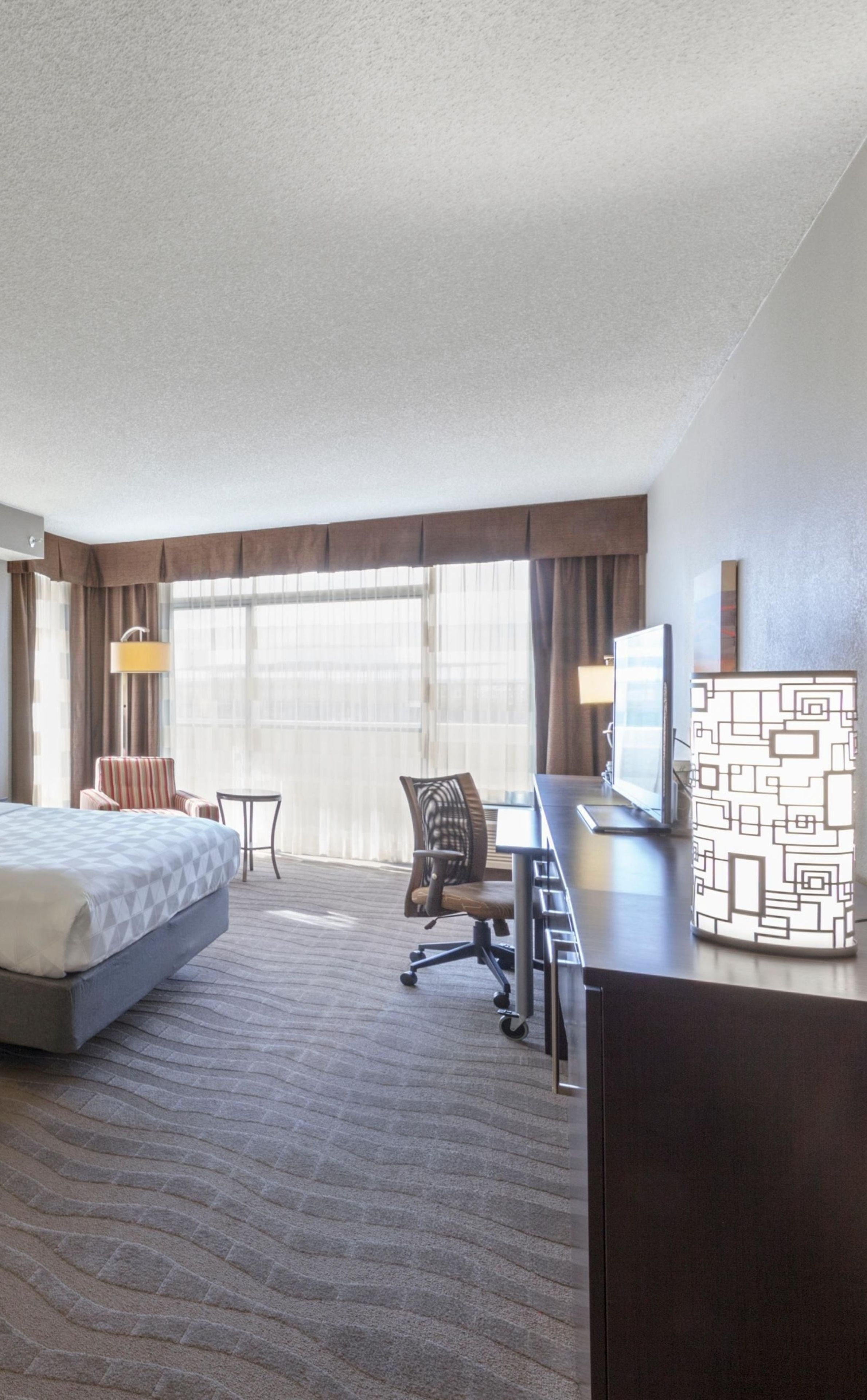 A hotel room with a bed, desk, chair, lamp, and a large window.