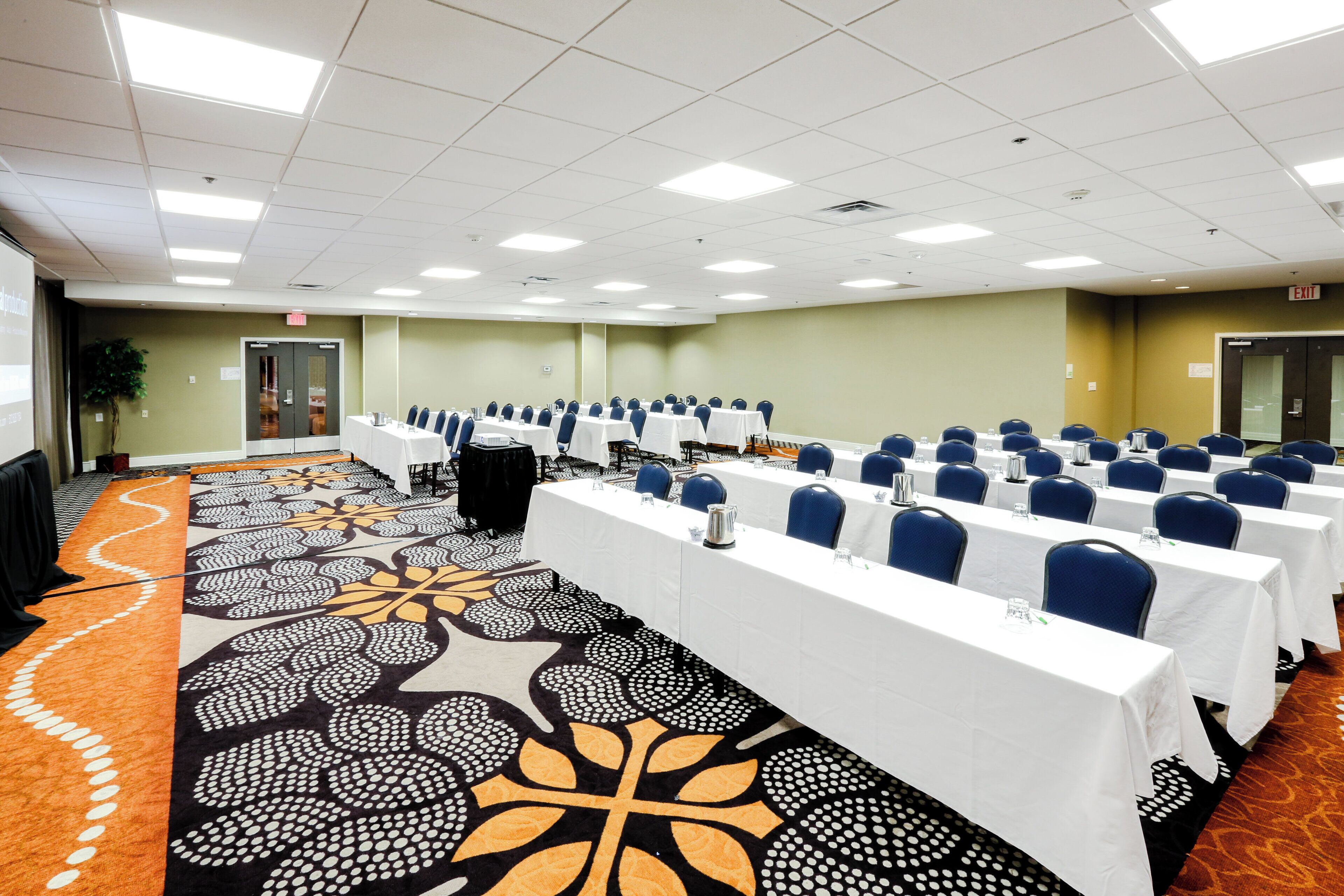 A conference room with rows of tables and chairs, a patterned carpet, and a plain wall with an exit sign.