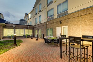 Een terras buiten met tegels, zitplaatsen en een gebouw met veel ramen en een bakstenen gevel.