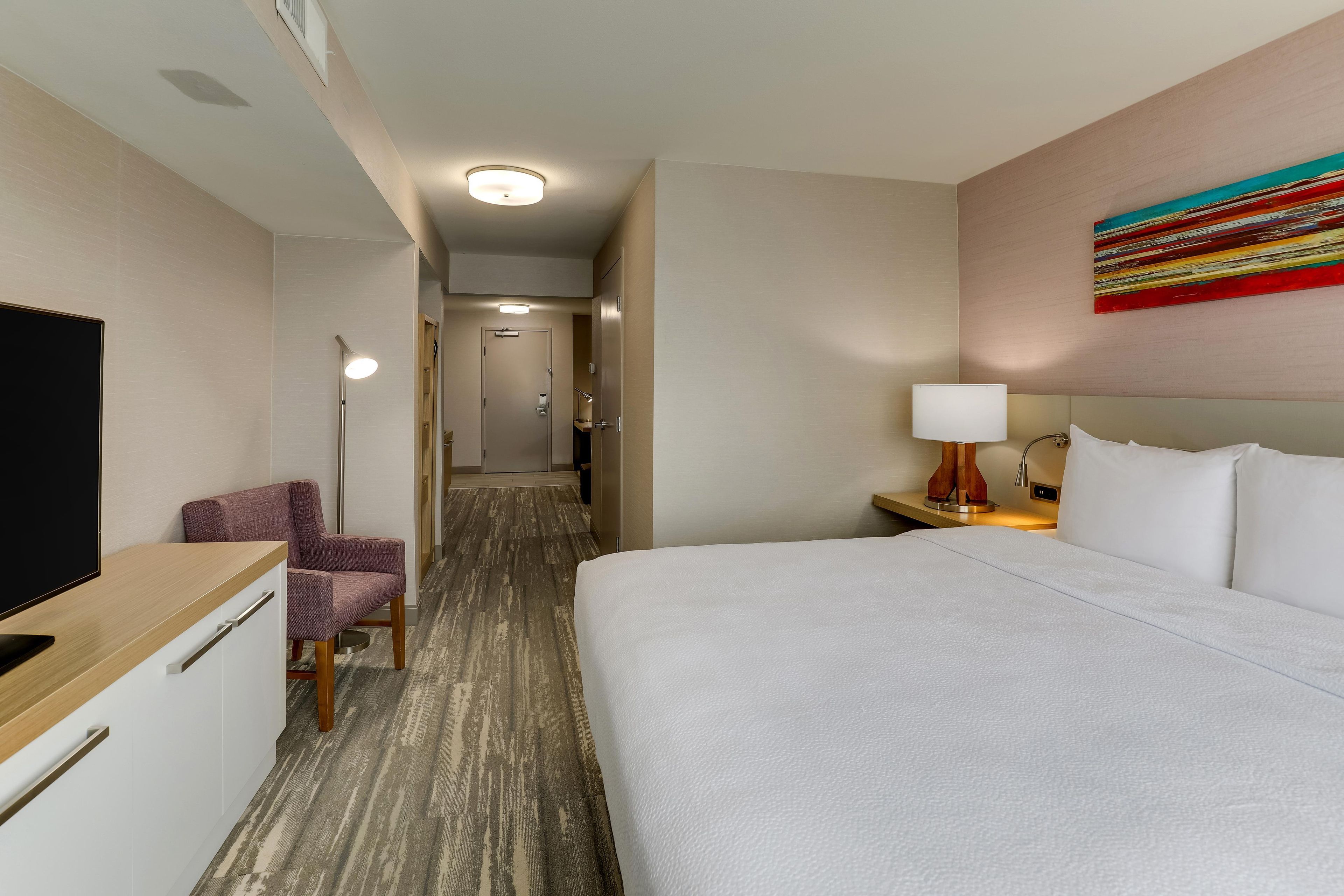Una habitación de hotel con una cama, un escritorio con televisor, una silla, una mesita de noche con lámpara, y un pasillo que conduce a otra habitación.