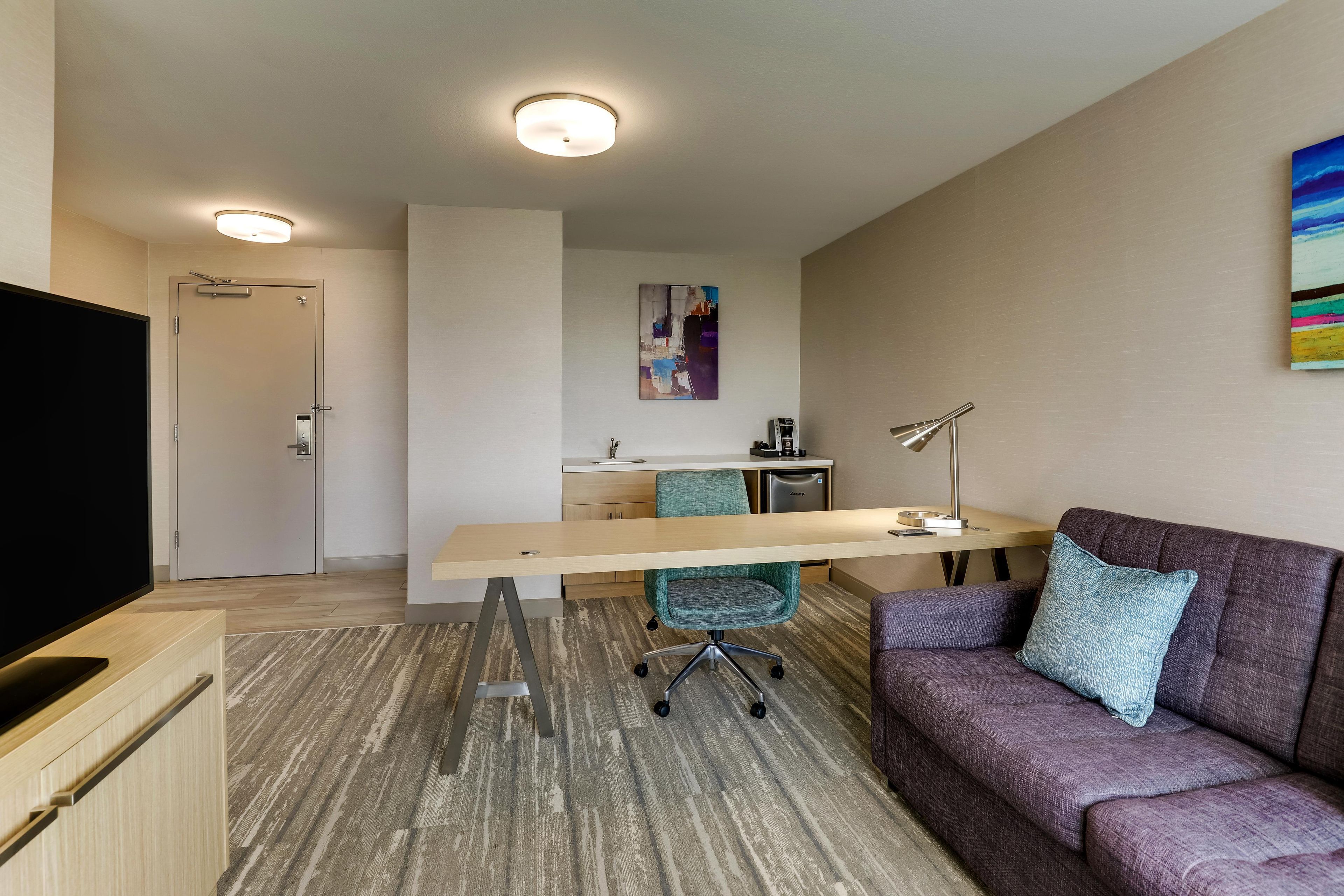 Una habitación de hotel moderna con televisión de pantalla plana, un escritorio con silla, un sofá y un cuadro en la pared.