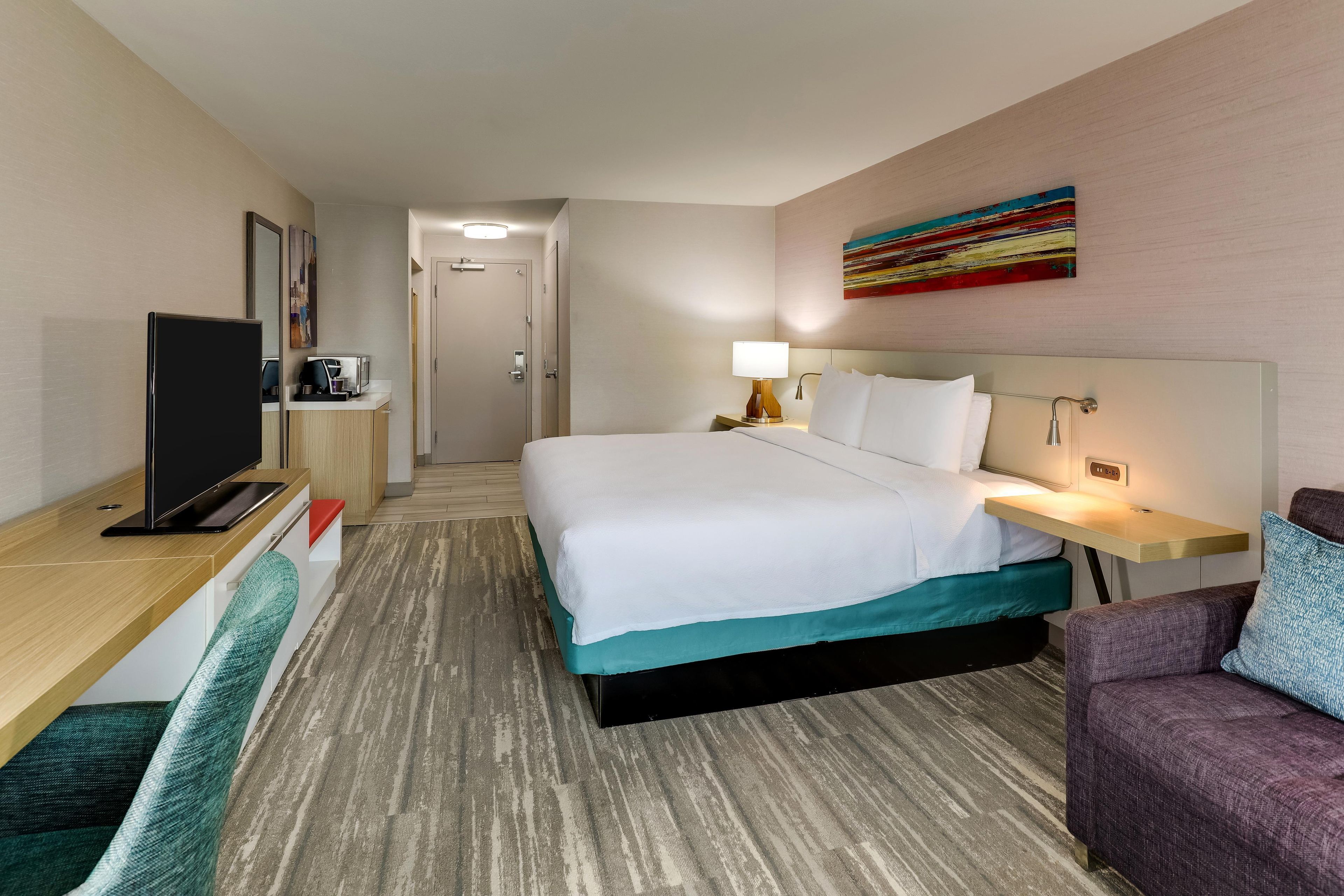 A hotel room with a bed, a desk with a flat-screen TV, a bedside table with a lamp, and a colorful abstract painting on the wall.