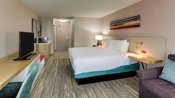 A hotel room with a bed, a desk with a flat-screen TV, a bedside table with a lamp, and a colorful abstract painting on the wall.