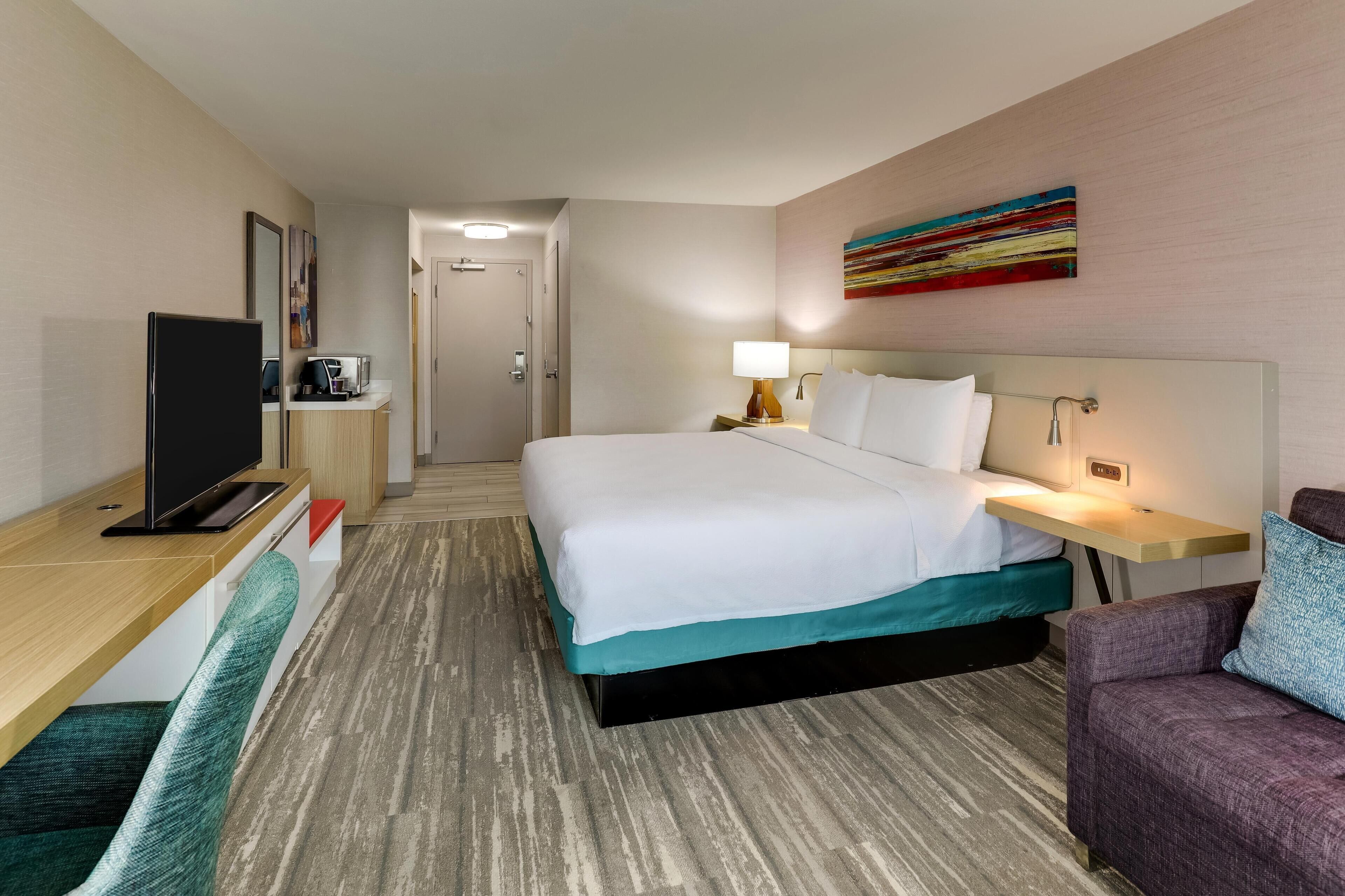 Une chambre d’hôtel comprenant un lit, un bureau avec une télévision, une chaise, une table de chevet avec une lampe, et une peinture abstraite aux couleurs vives accrochée au mur.