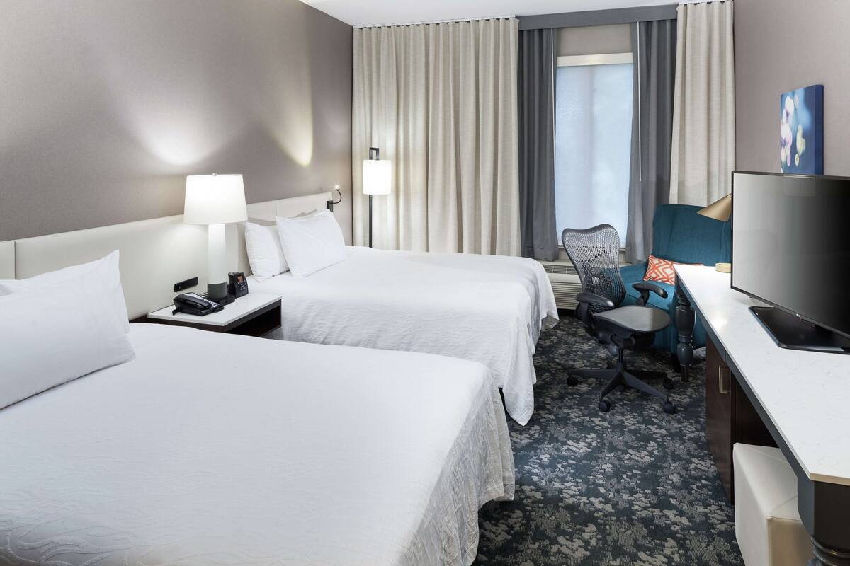 Une chambre d’hôtel avec deux lits, un bureau, une chaise, une télévision et une fenêtre avec des rideaux.