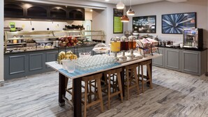 A buffet setup with a variety of food items and beverages, a long table with stools, and a refrigerator.