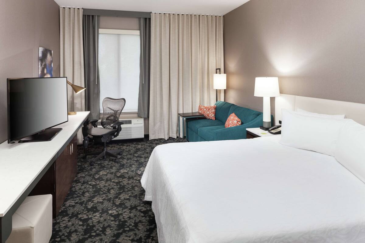 Une chambre d’hôtel avec un lit, un bureau avec une télévision, une chaise, un canapé et une fenêtre avec des rideaux.