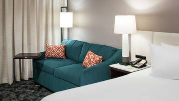 A hotel room with a teal sofa, two patterned pillows, a bedside table with a lamp, a phone, and a bed with white linens.