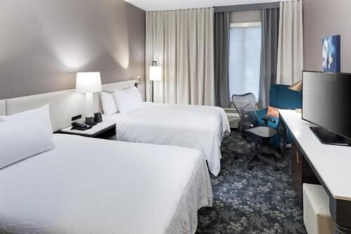 Une chambre d’hôtel avec deux lits, un bureau, une chaise, une lampe et une fenêtre avec des rideaux.