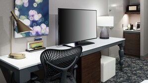 A hotel room with a desk, a flat-screen TV, a lamp, a chair, a microwave, and a small kitchenette.